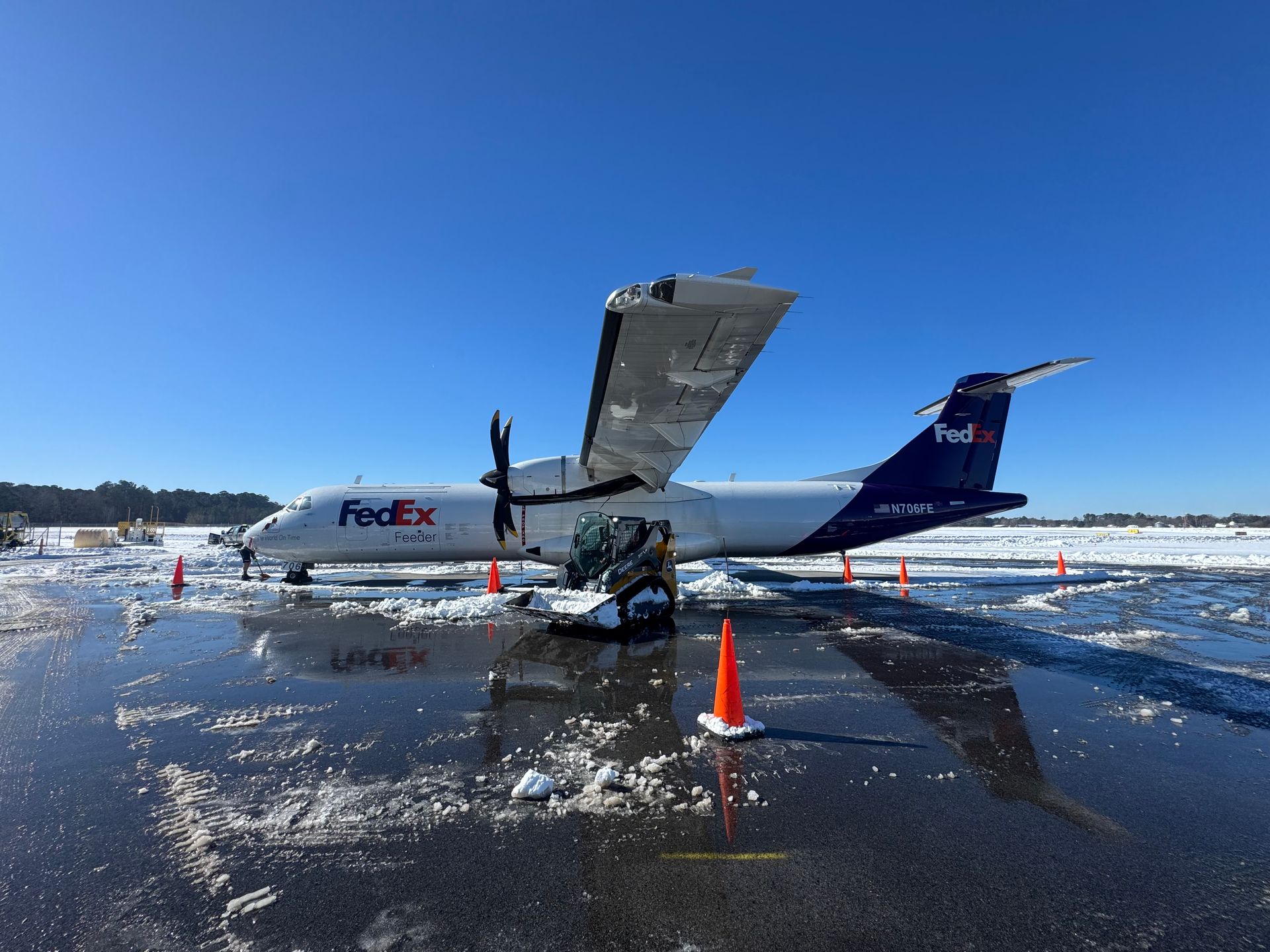 A FedEx cargo plane parked on a wet, snow-covered tarmac with orange safety cones nearby under a clear blue sky.