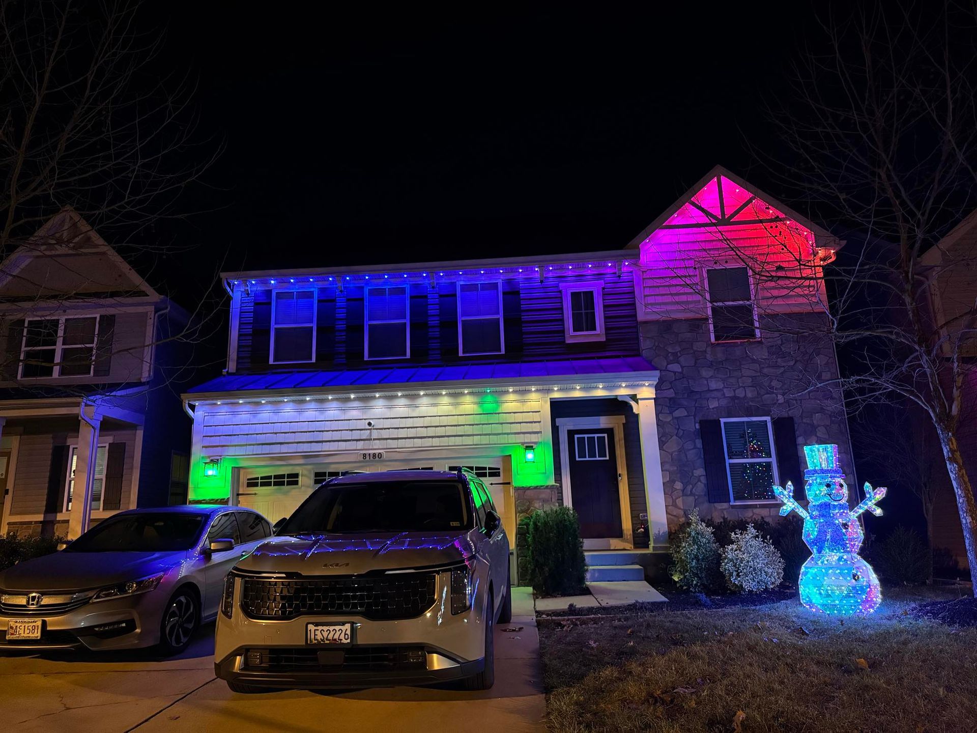 A two-story house at night with festive blue and magenta outdoor lights and a glowing snowman figure on the front lawn.