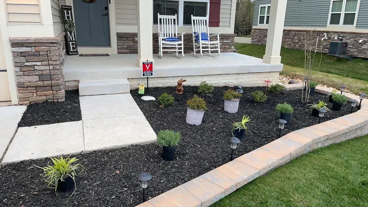 A front porch with white rocking chairs and a landscaped garden featuring dark mulch, small potted plants, and stone edging.
