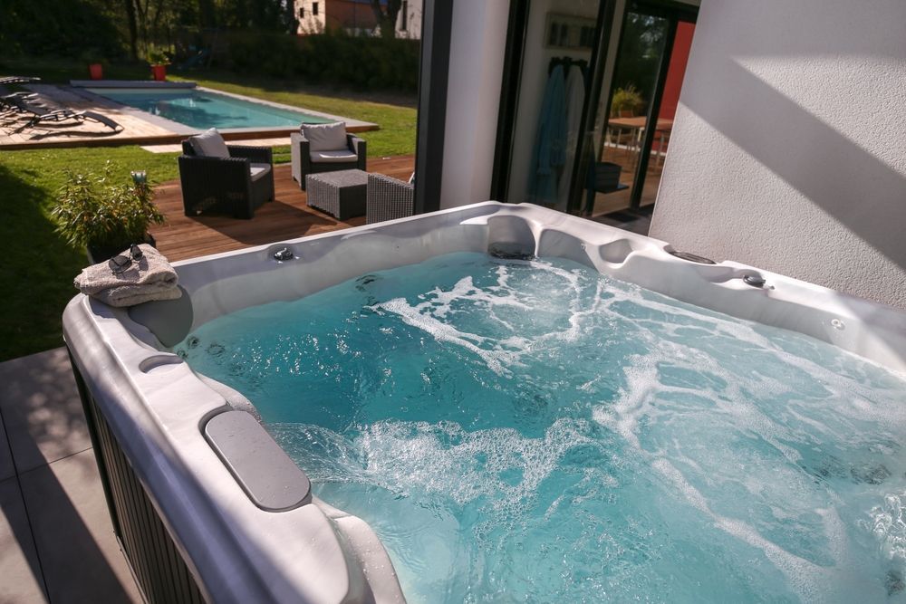 A white hot tub with bubbling water next to a patio with lounge furniture and a swimming pool in the background.