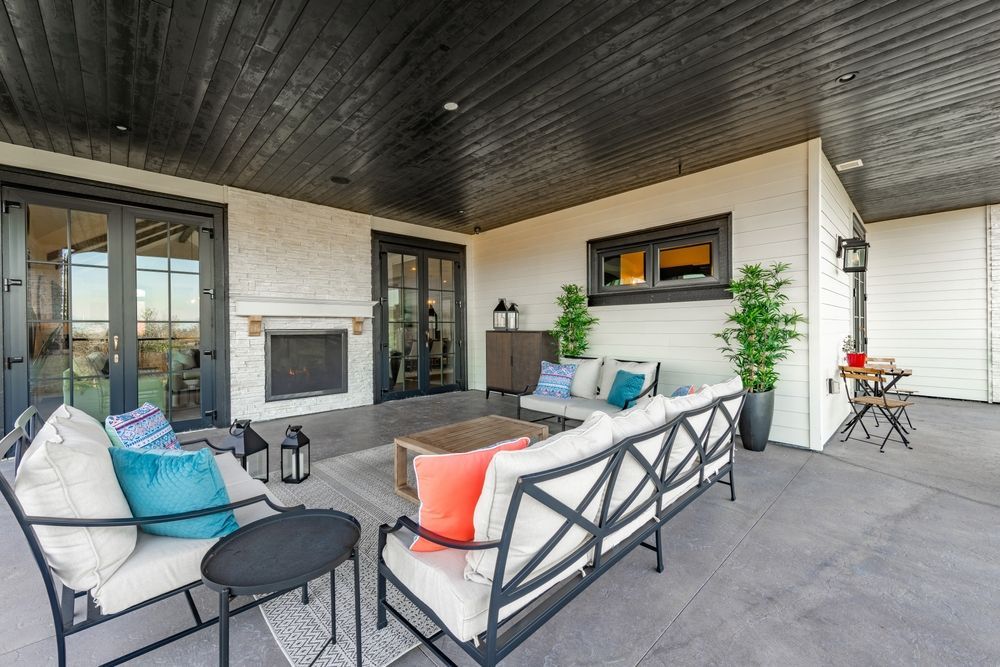 A covered outdoor patio featuring a fireplace, modern patio furniture with colorful pillows, and a black wood ceiling.