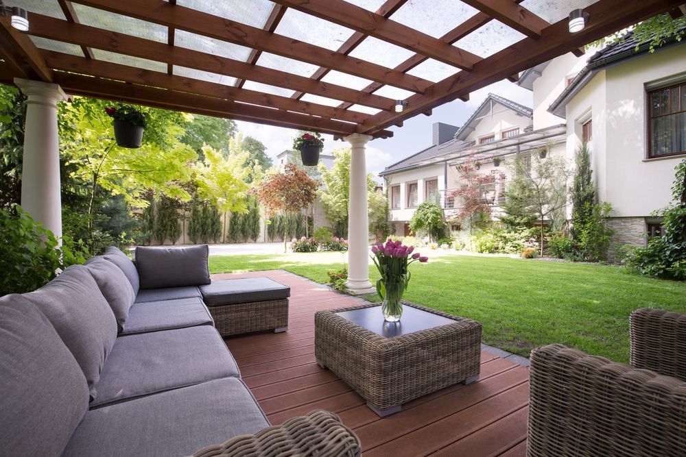 A cozy outdoor patio with grey wicker furniture and a wooden pergola overlooking a lush green garden and house.
