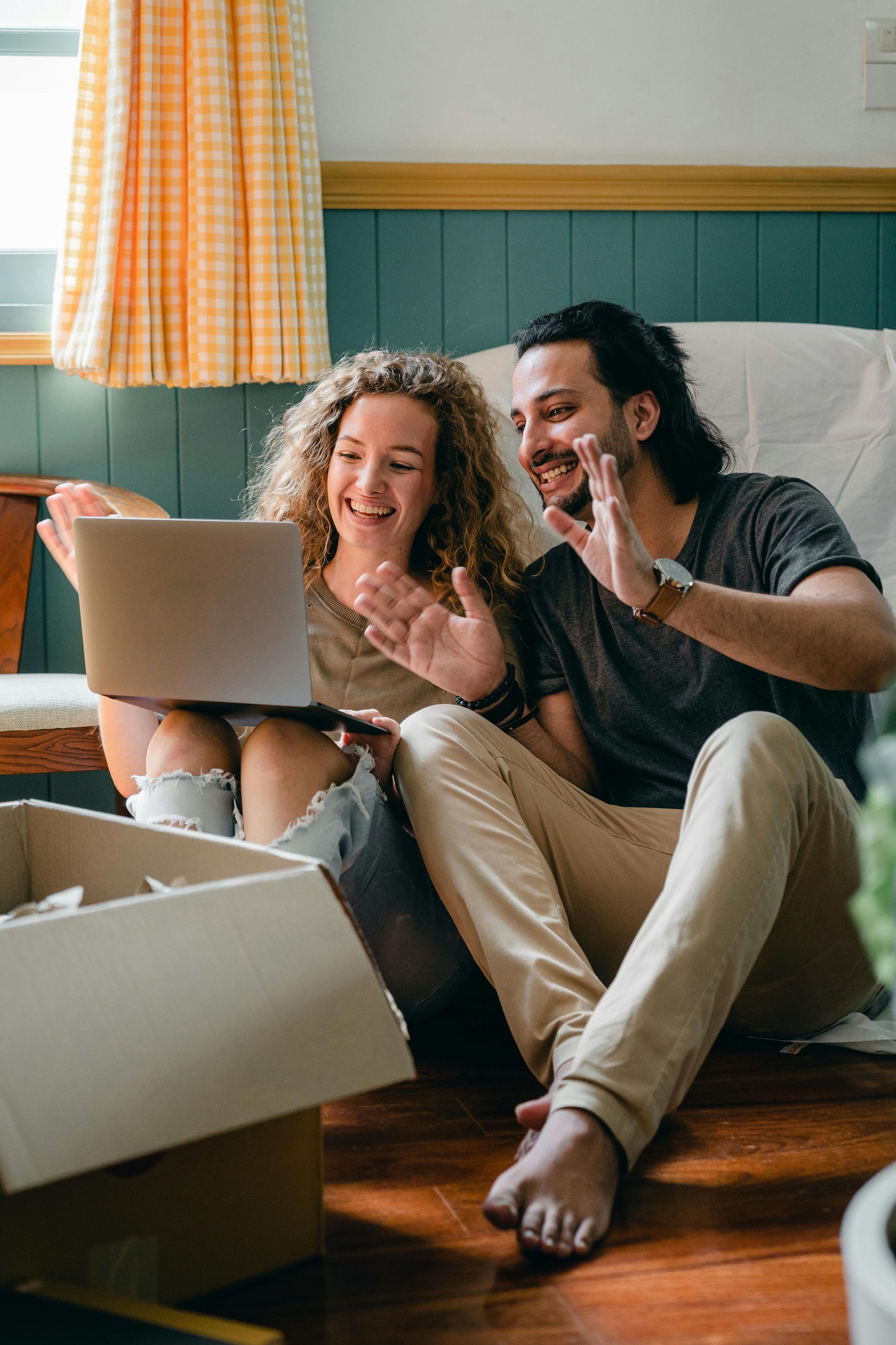 Couple on a wooden floor, waving at a laptop screen. Smiling, inside, boxes nearby.
