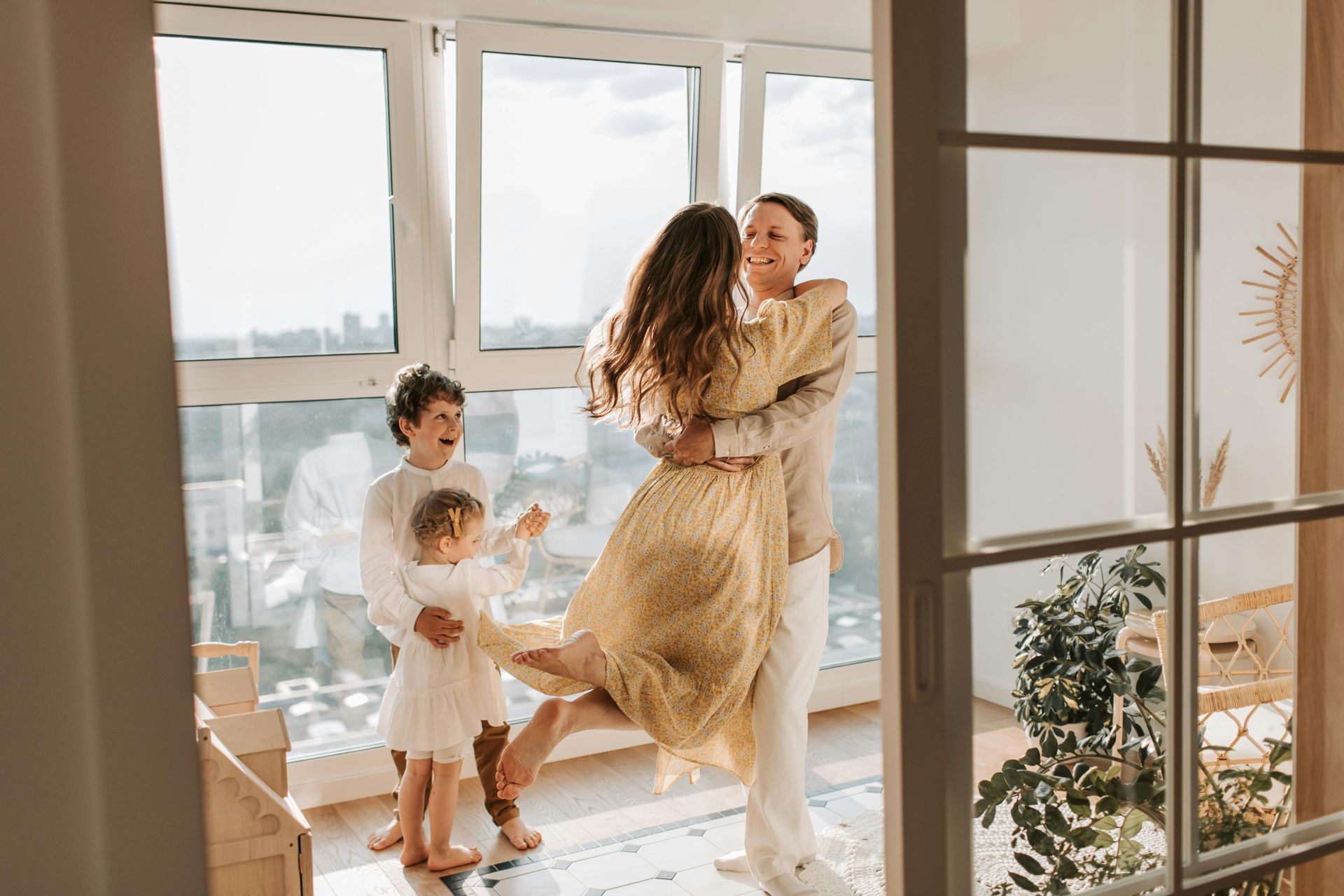 Family of four hugging near large windows, the mother being lifted, children laughing.