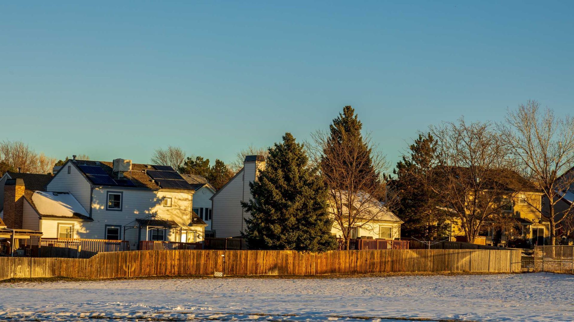 A snowy field with a fence and houses in the background.