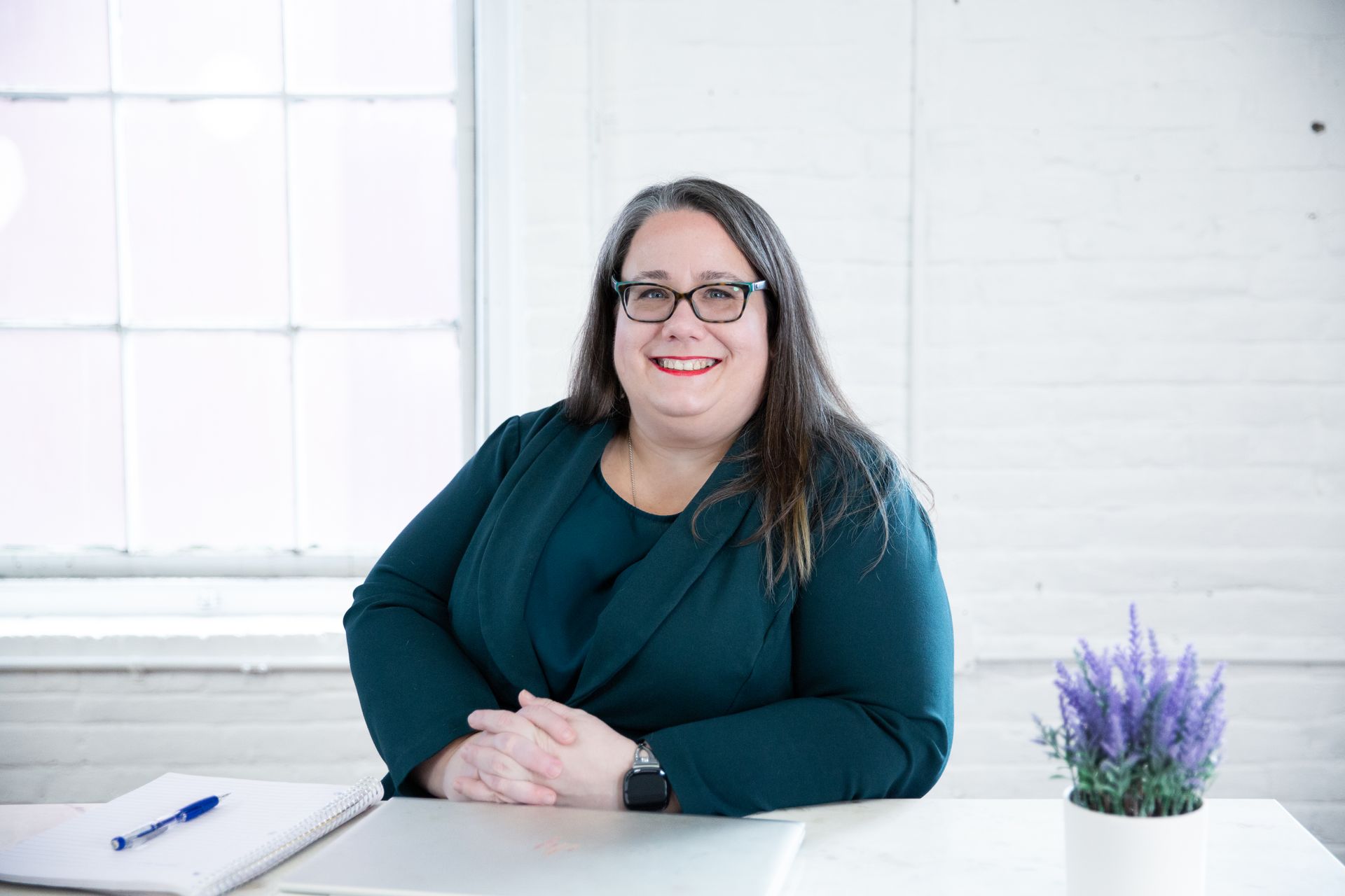 A woman is sitting at a desk with her hands folded and smiling.