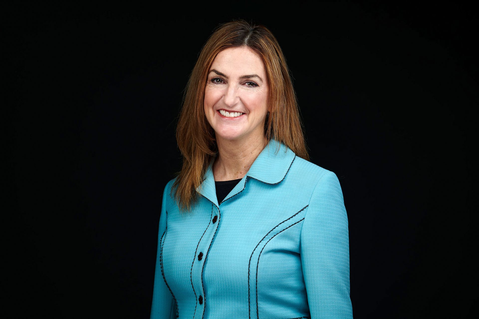 A woman in a blue jacket is smiling in front of a black background.