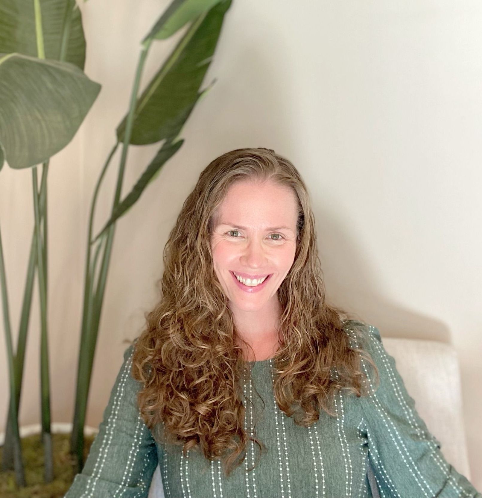 A woman is smiling while sitting on a couch in front of a plant.