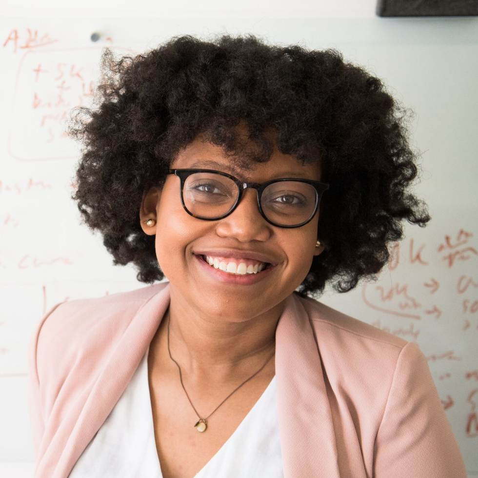 A woman wearing glasses and a pink jacket smiles in front of a white board