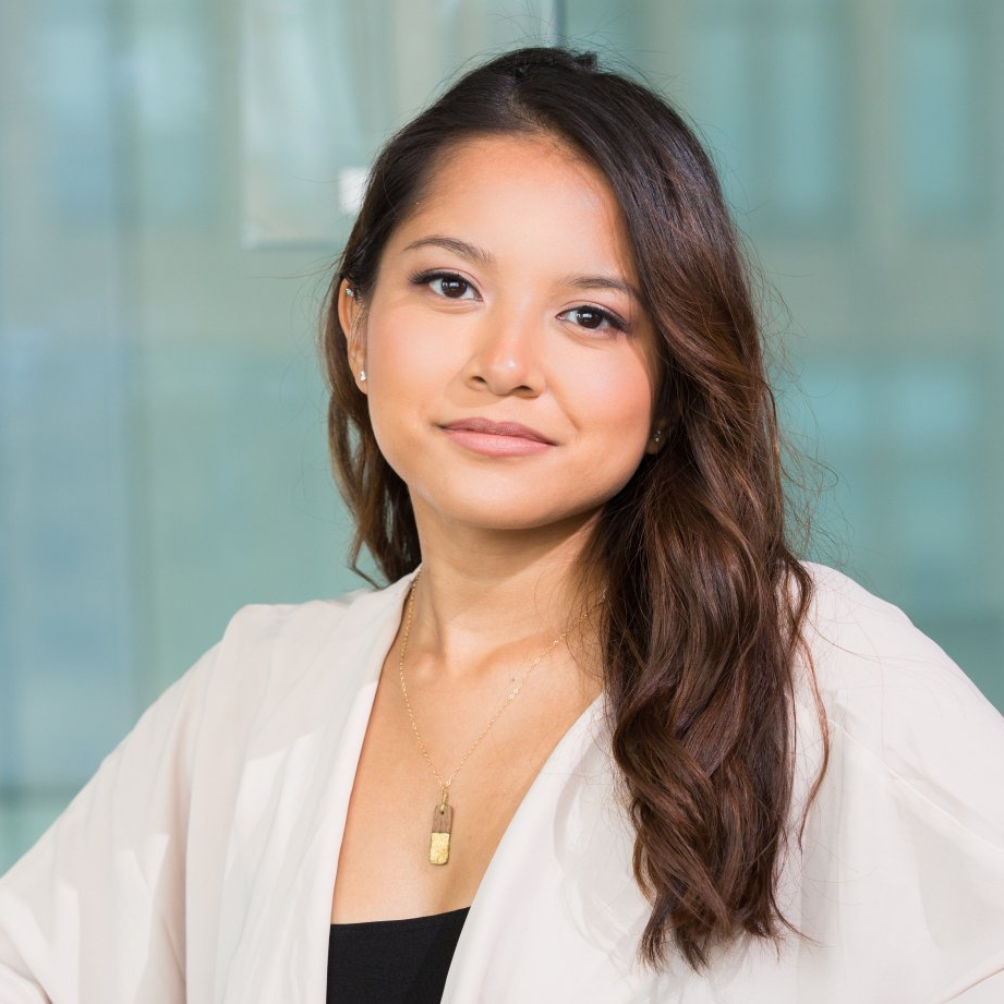 A woman wearing a white jacket and a necklace is standing in front of a glass wall.