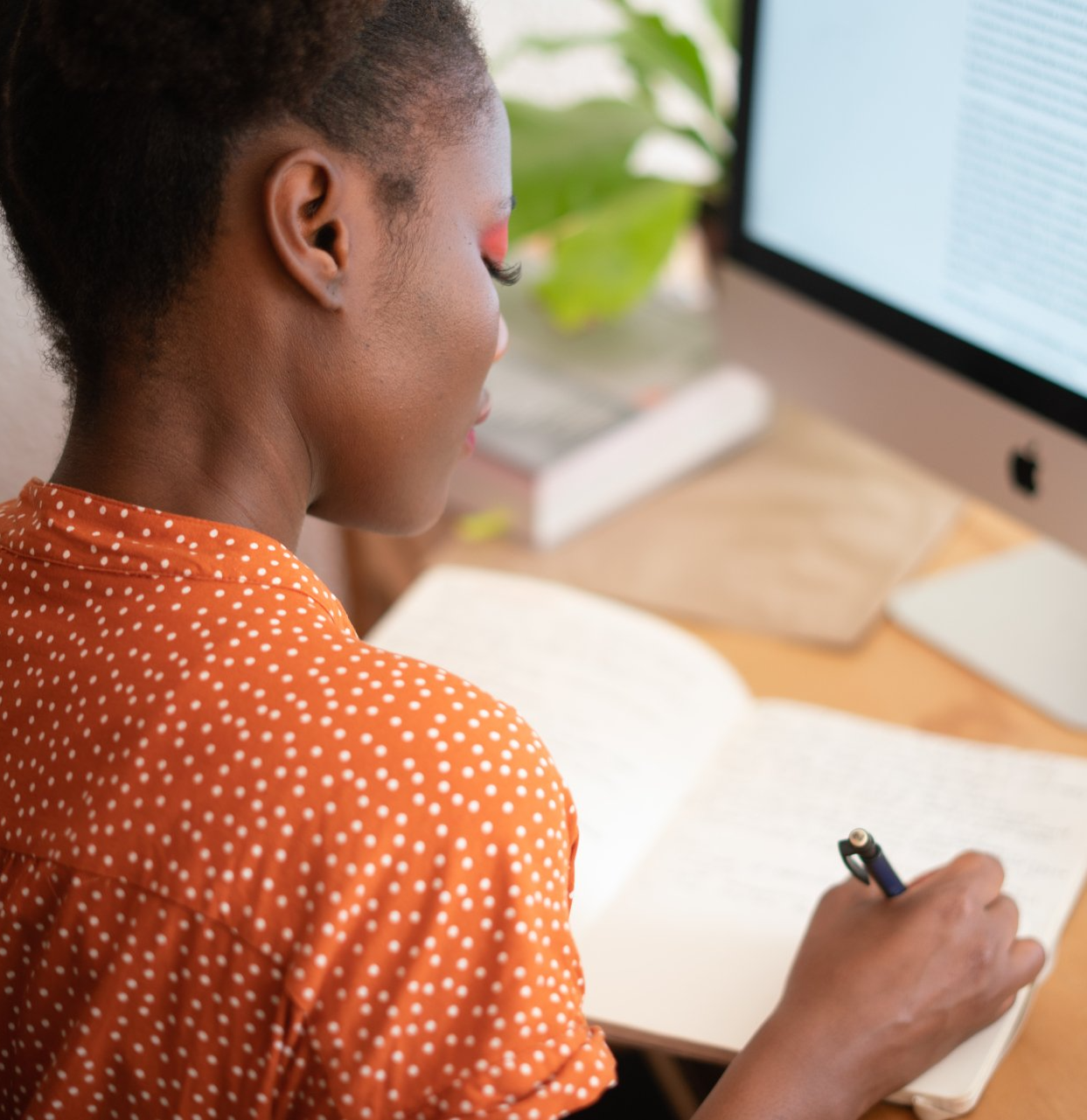 A woman is writing in a notebook while looking at a computer screen