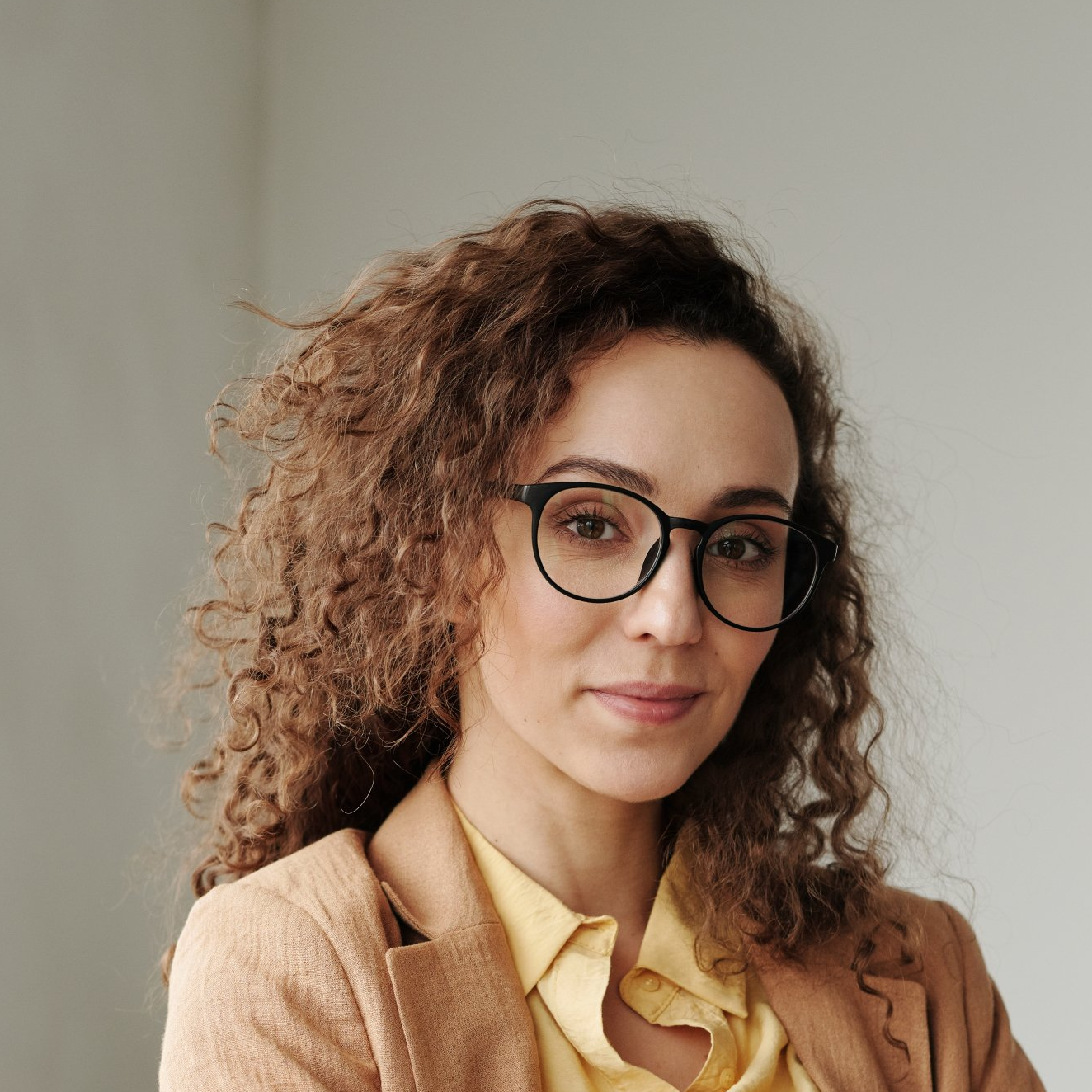 A woman with curly hair and glasses is wearing a tan jacket and a yellow shirt.