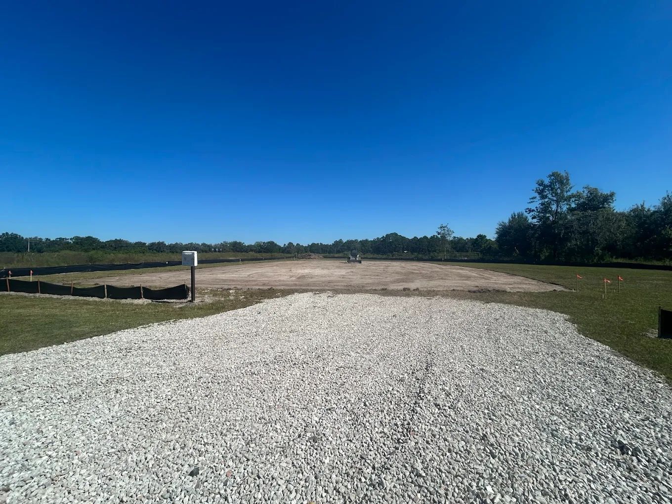 A gravel driveway leads to a cleared, sandy construction lot under a clear blue sky, bordered by black silt fencing.