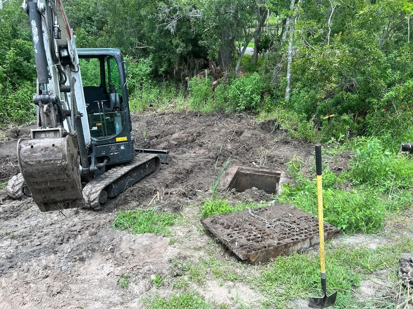 A mini excavator sits on a dirt plot beside an open concrete septic tank lid and a shovel in a wooded area.
