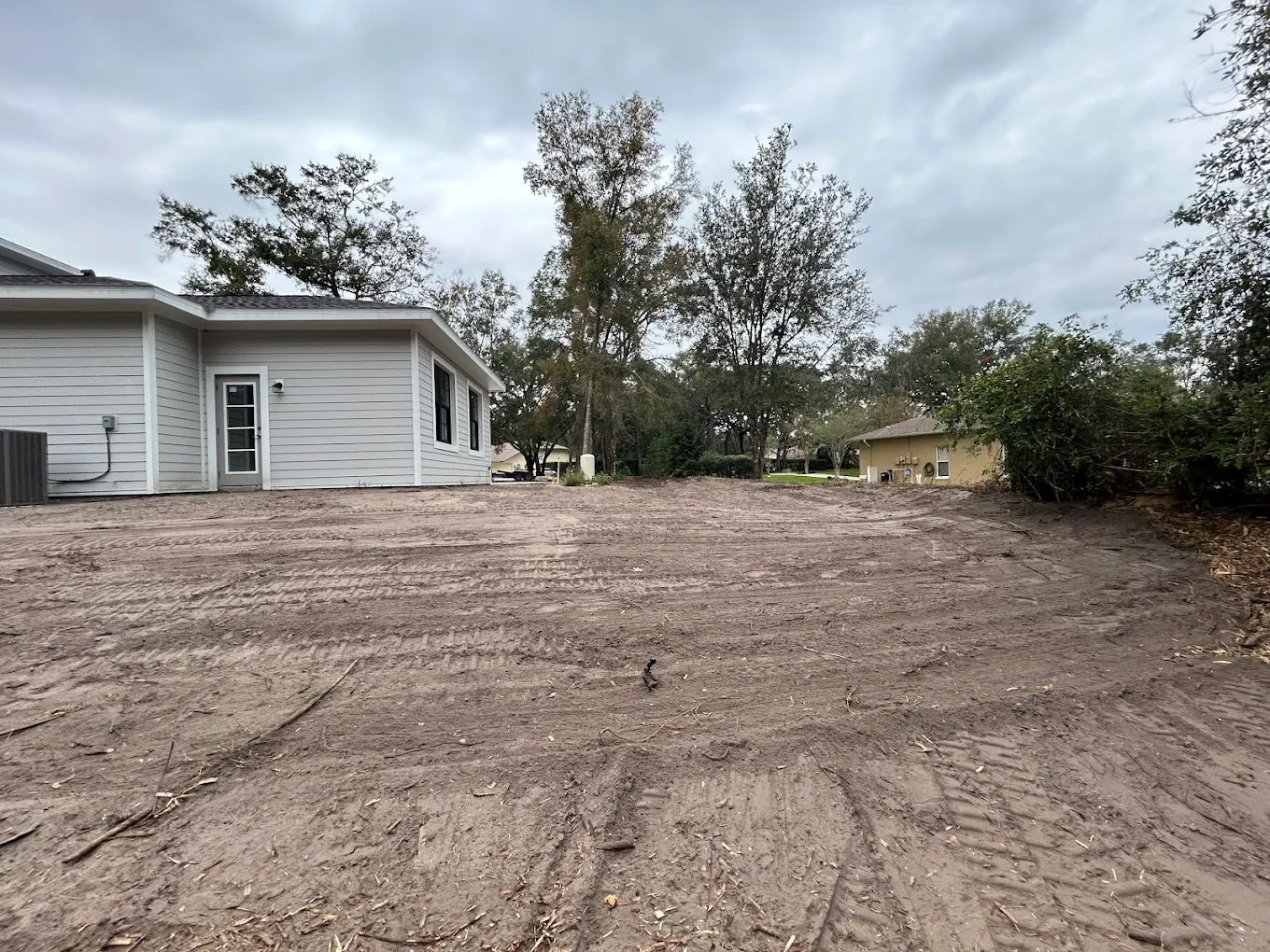 A light-gray house sits next to an expansive, unlandscaped dirt yard under a cloudy sky.