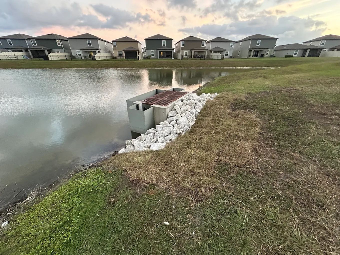 A concrete drainage structure at the edge of a pond, surrounded by white riprap rocks, with houses in the background.