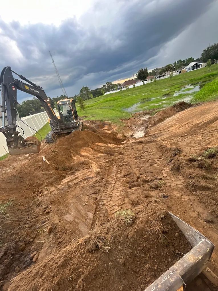 A dark excavator sits on a dirt mound at a construction site under a cloudy sky near a grassy, flooded field.