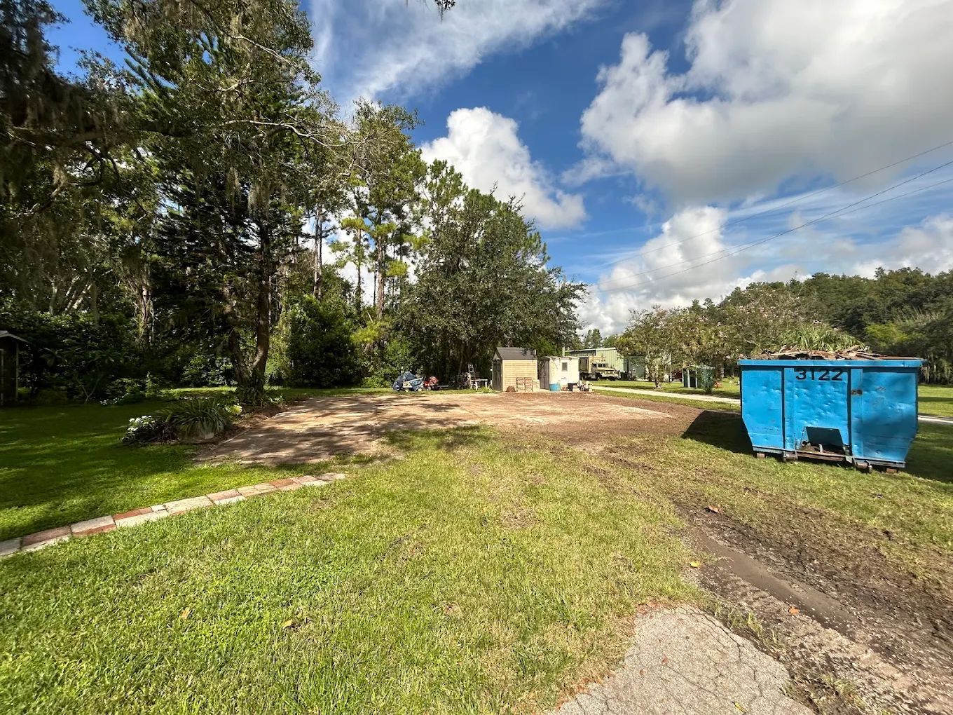 A vacant, dirt-covered lot next to a blue dumpster on a sunny day with trees in the background.