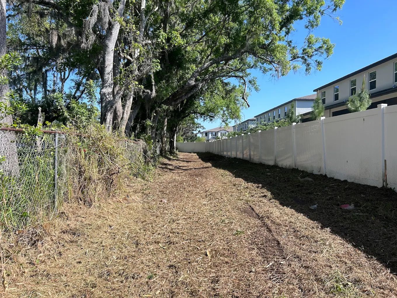 A path covered in wood mulch runs between a chain-link fence with trees on the left and a white vinyl fence on the right.