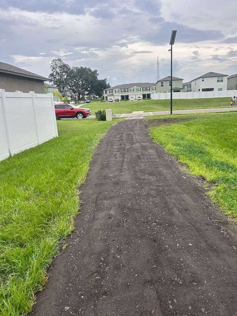 A dirt path leads through a grassy residential area, bordered by a white fence on the left and a street in the distance.