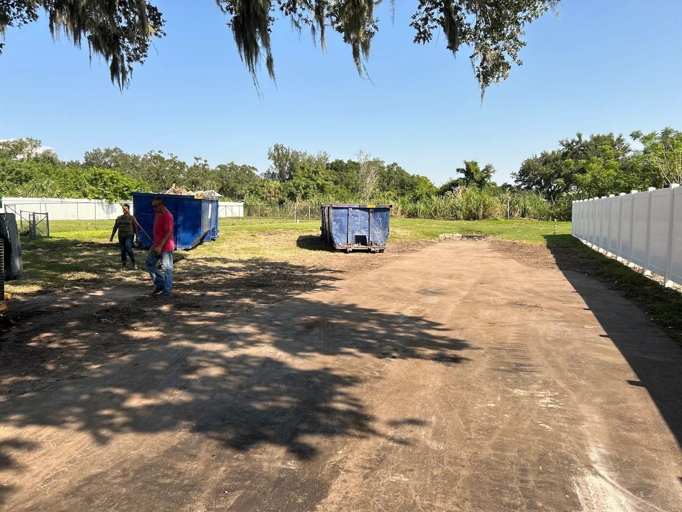 Two people clear debris near two blue dumpsters on a dirt lot next to a white fence on a sunny day.