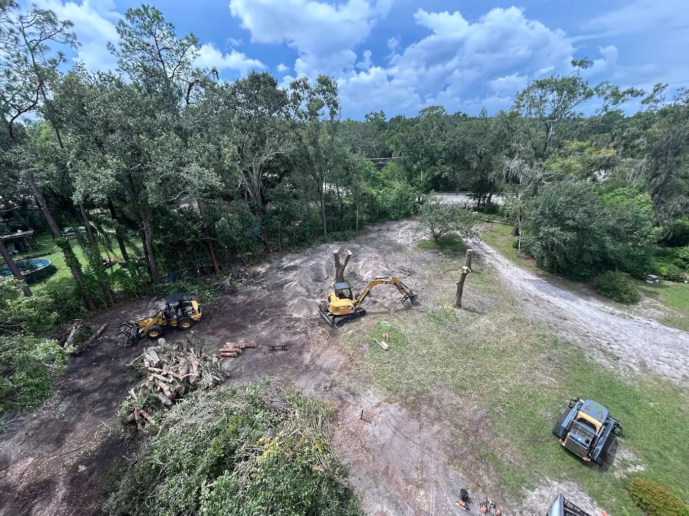 An aerial view of a wooded lot being cleared by yellow construction equipment and excavators on a sunny day.
