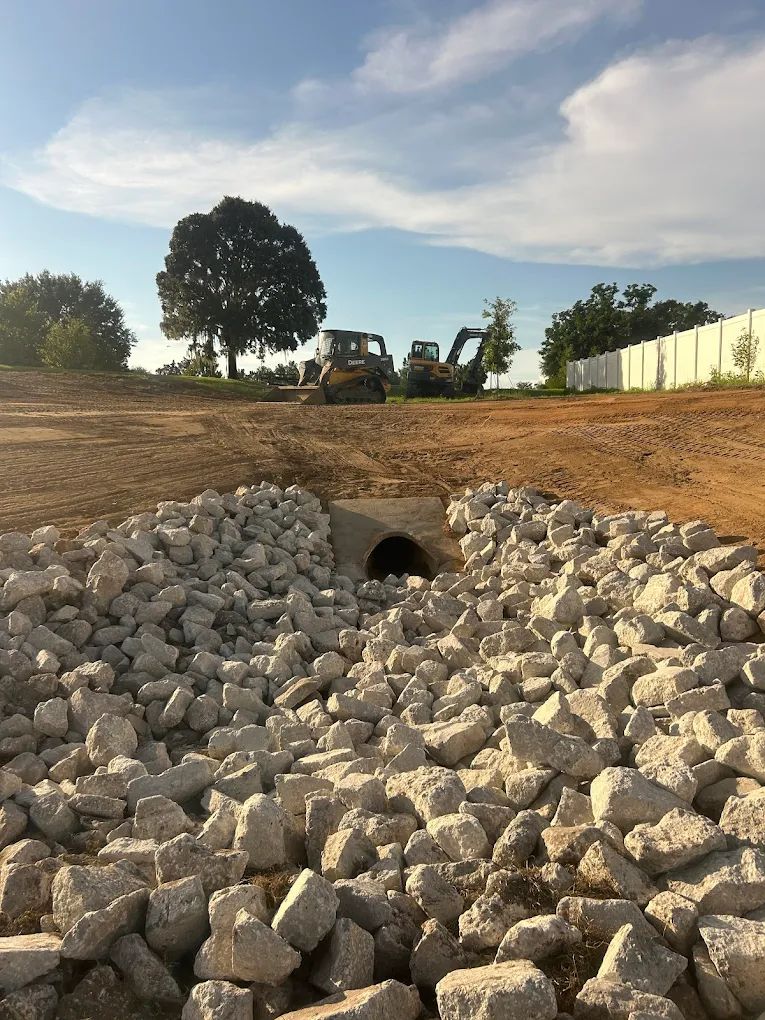 A concrete drainage pipe surrounded by riprap stone in a construction site with heavy machinery in the background.