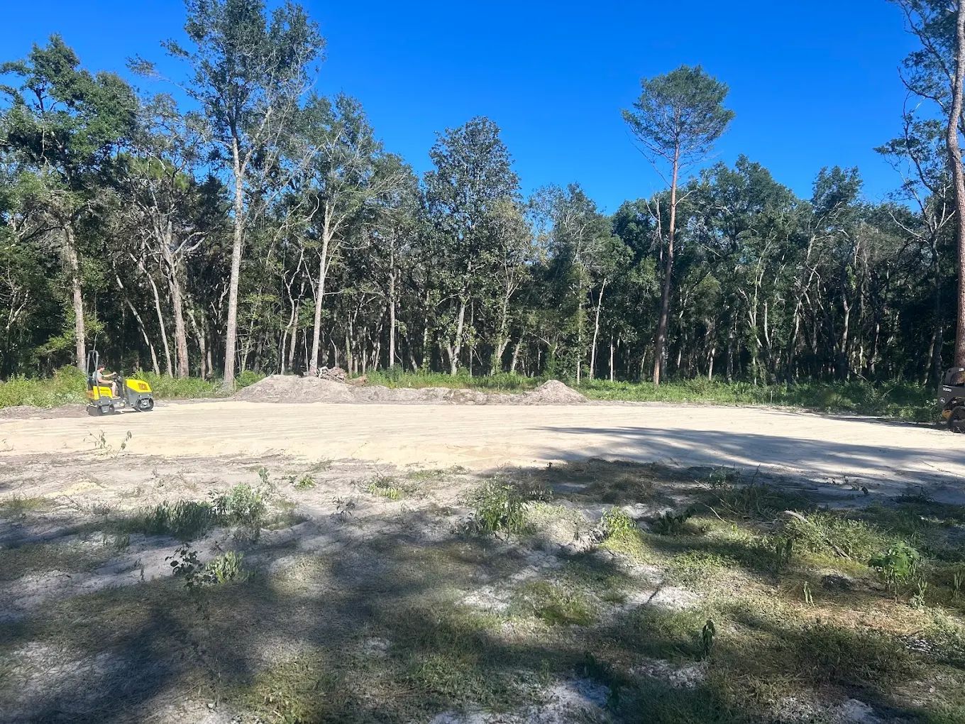 A clearing with light-colored soil and a yellow vehicle near a dense forest line under a clear blue sky.