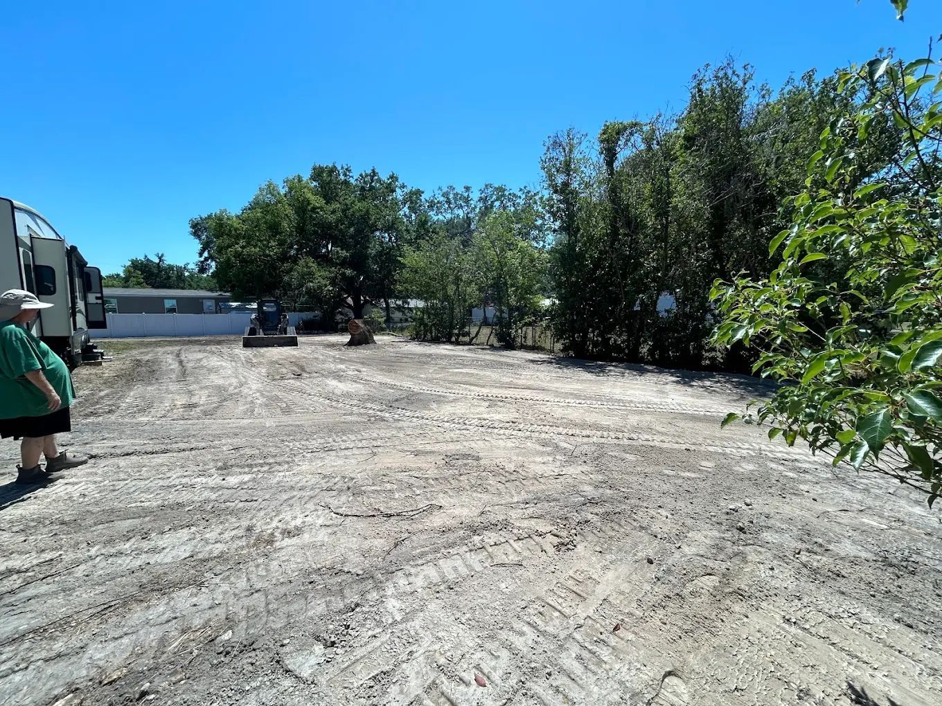 A person stands on a large, empty dirt lot next to an RV, with trees and a mobile home in the background.