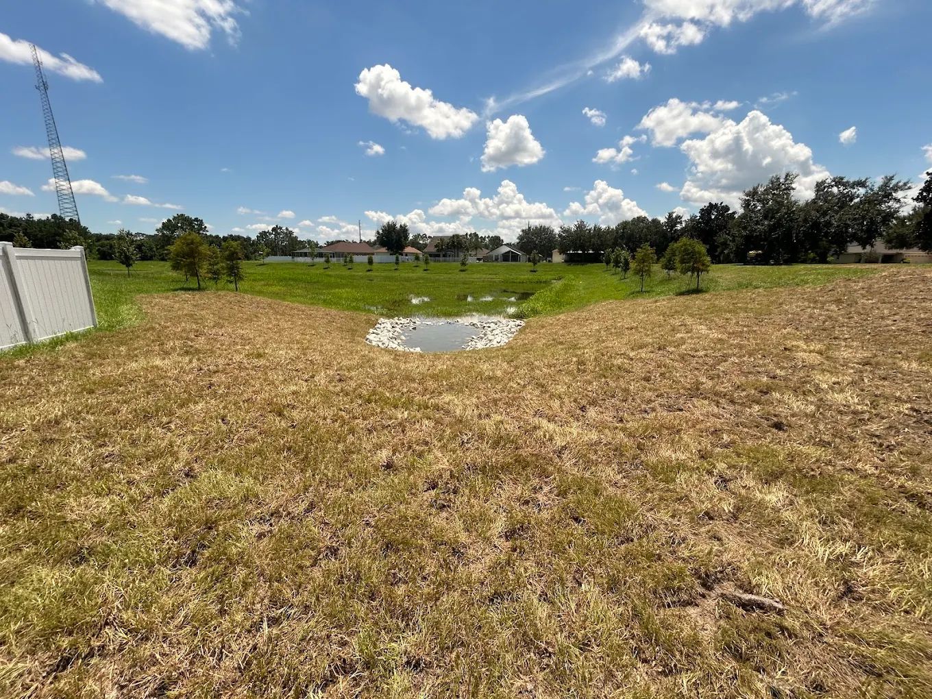A sloped, grassy field leads down to a small, rock-lined drainage pond under a blue sky with scattered white clouds.
