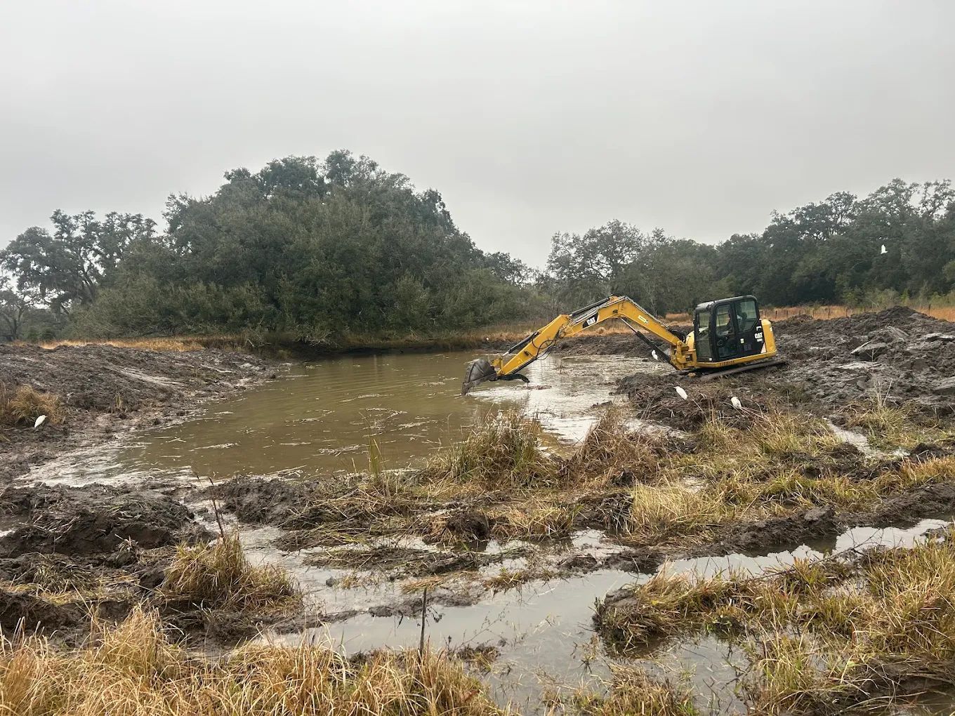 A yellow excavator works in a muddy, water-filled clearing near a dense treeline on an overcast day.