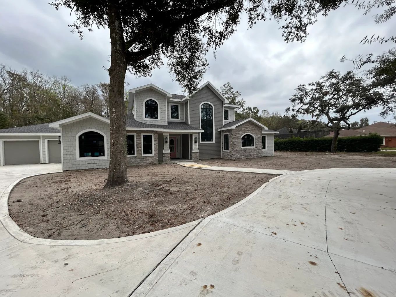 A two-story grey house with a stone facade, a concrete driveway, and a large tree in the front yard.
