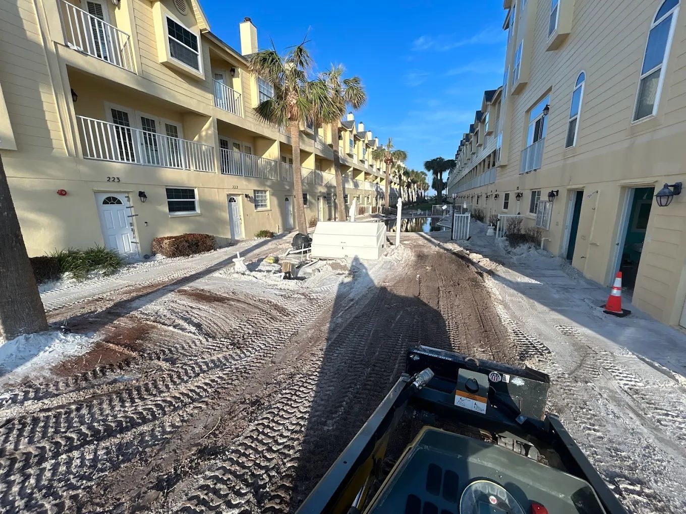 Construction equipment tracks on a dirt ground between two apartment buildings on a sunny day.