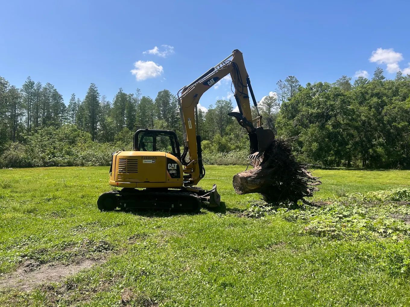 A yellow Caterpillar excavator in a grassy field, lifting a large tree stump with its bucket under a clear blue sky.