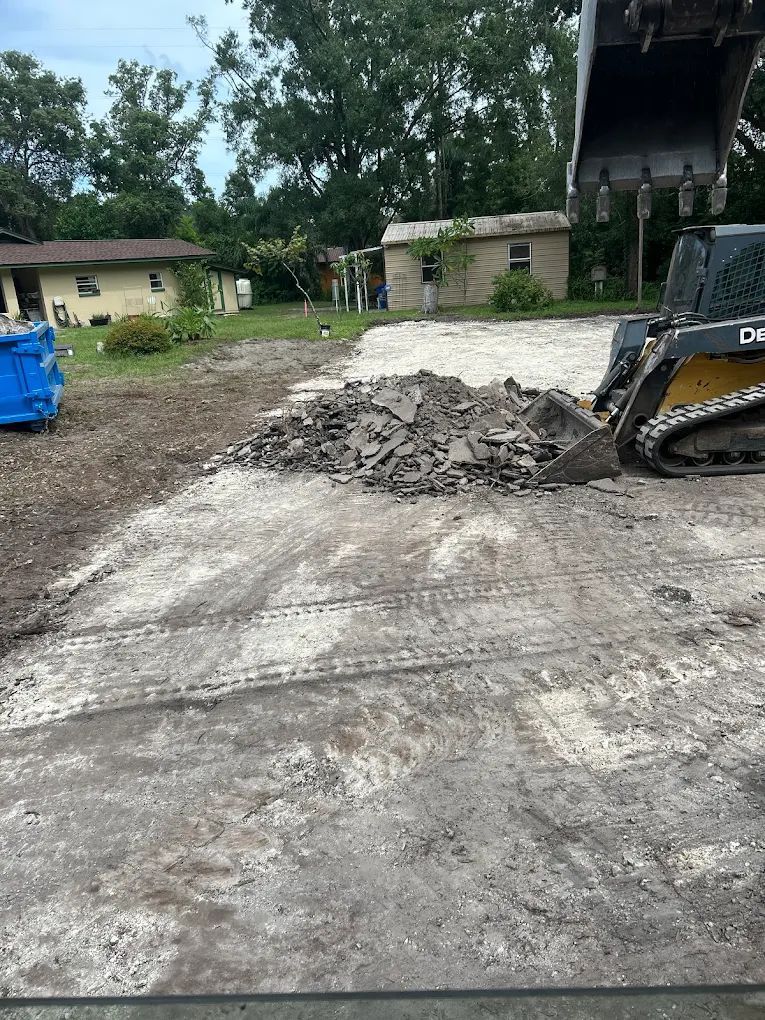 A skid steer loader sits on a gravel lot next to a pile of broken concrete, with houses visible in the background.