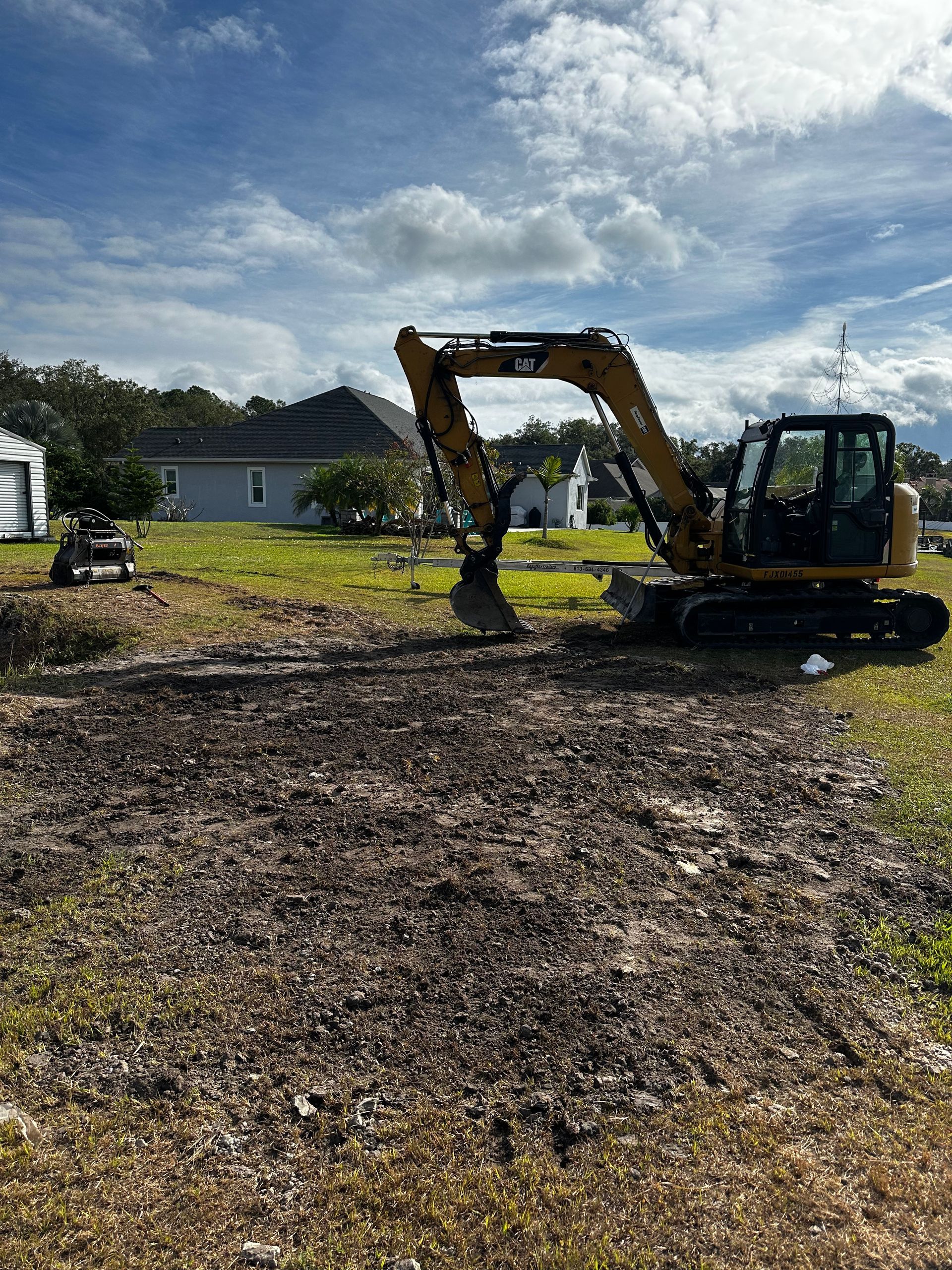 A yellow CAT excavator sits on a grassy field in front of a residential home under a blue, cloudy sky.
