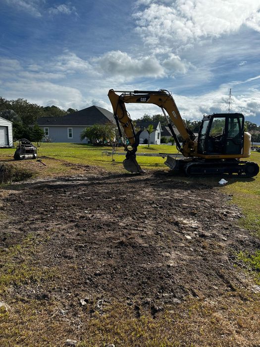 A yellow excavator is parked on a cleared, dark dirt patch in a residential backyard under a sunny, partly cloudy sky.