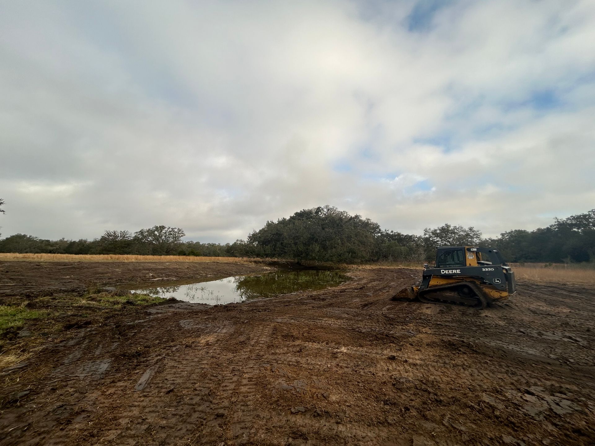 A construction bulldozer sits on muddy ground beside a small pond, with trees and a cloudy sky in the background.