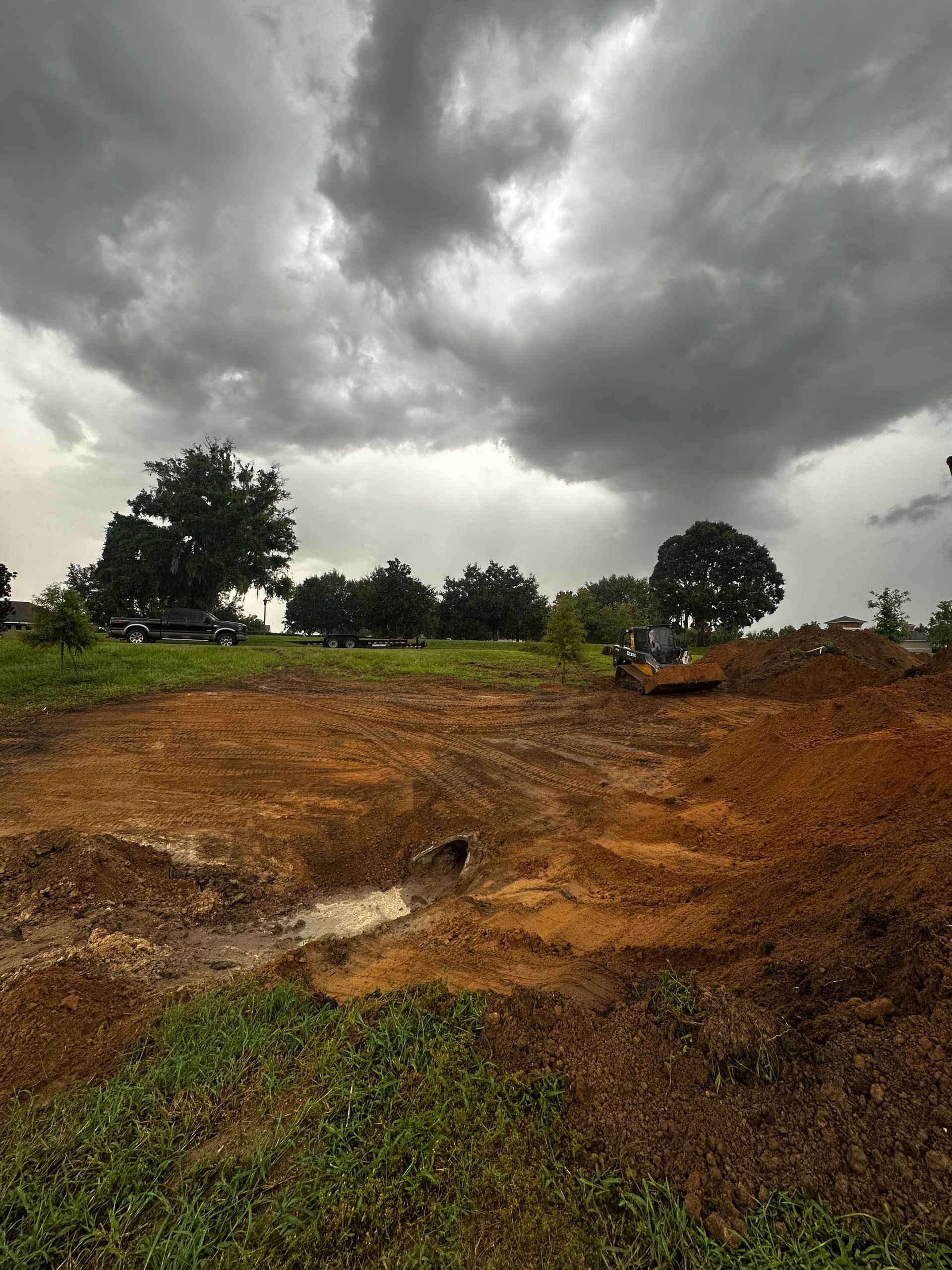 A construction vehicle excavates a muddy plot of land under a dark, stormy sky with trees in the background.