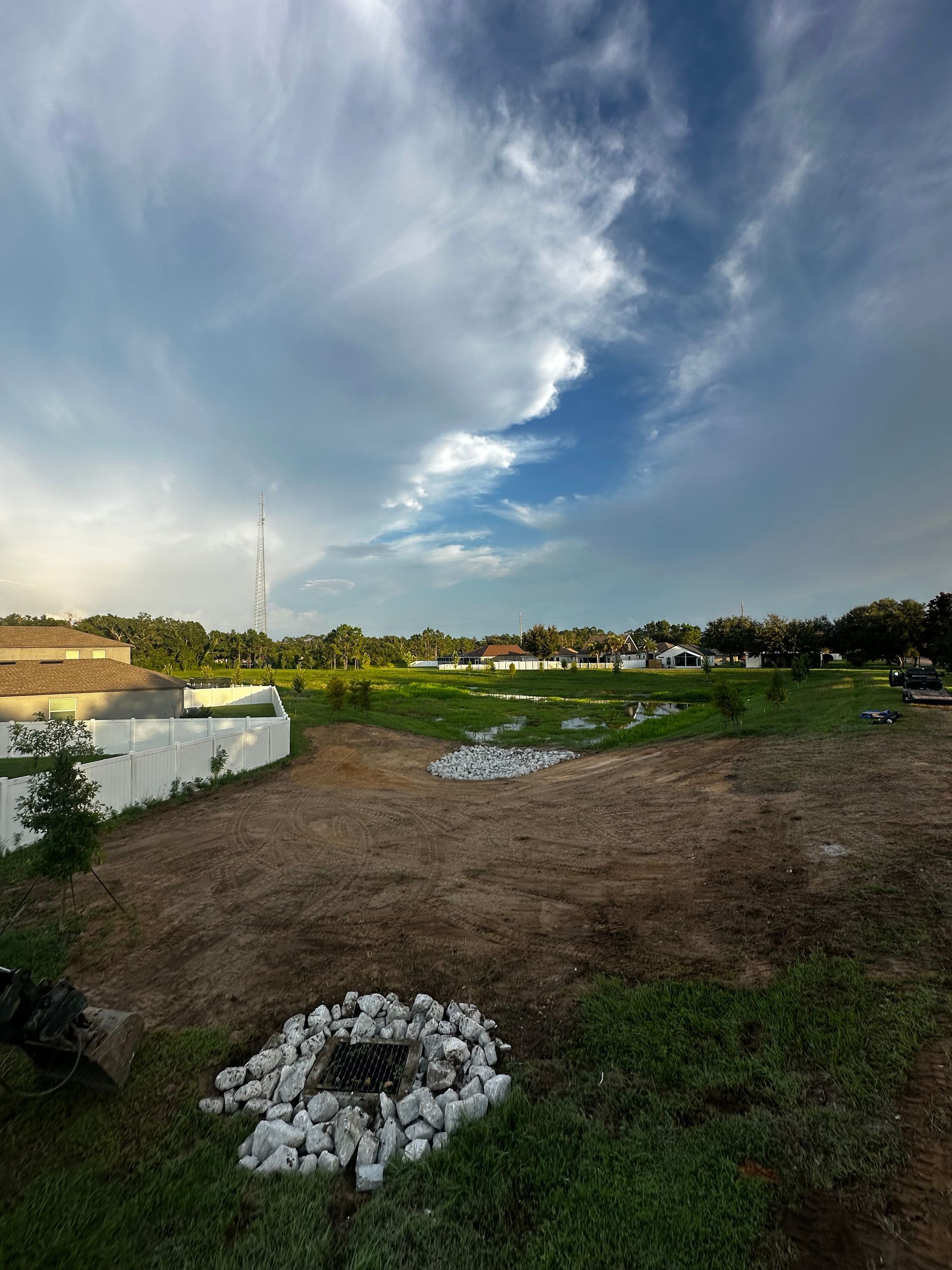 A grassy lot under a cloudy, blue sky with two circular, white rock drainage features in the foreground.
