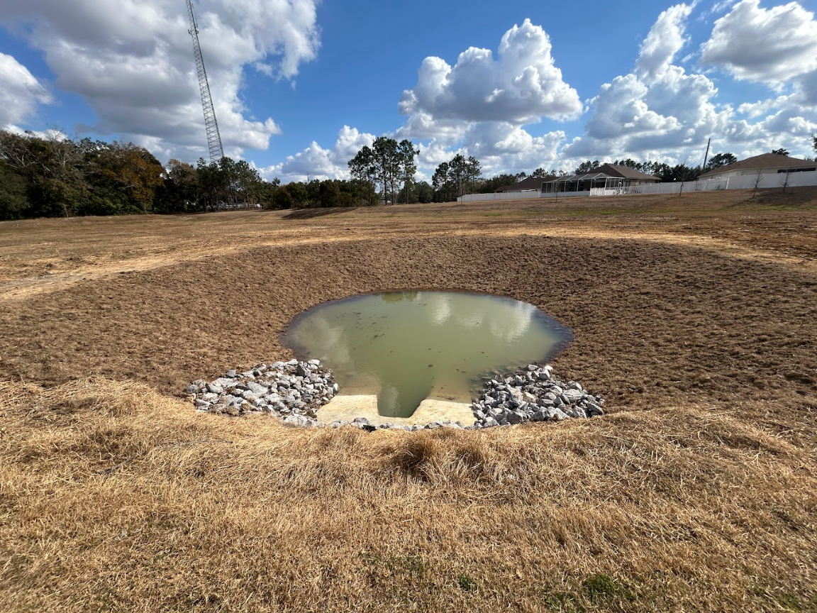 A small, circular retention pond with a stone-lined spillway sits in a grassy field under a bright, cloudy sky.