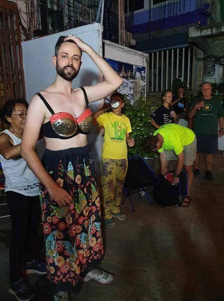 A man in a bra and floral skirt is standing in front of a group of people