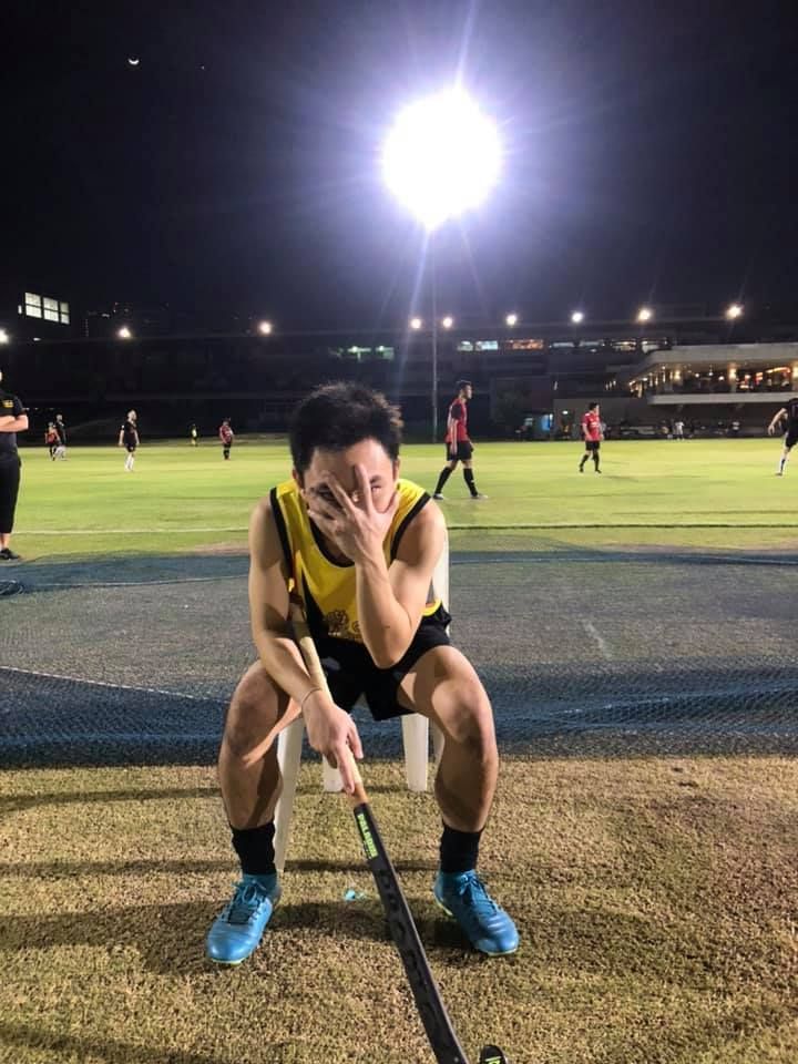 A man is sitting on a bench with a hockey stick in front of a field.