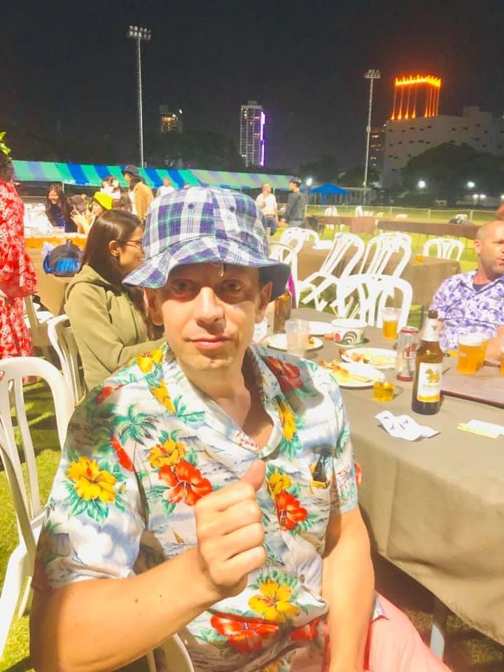 A man in a hawaiian shirt is giving a thumbs up while sitting at a table.