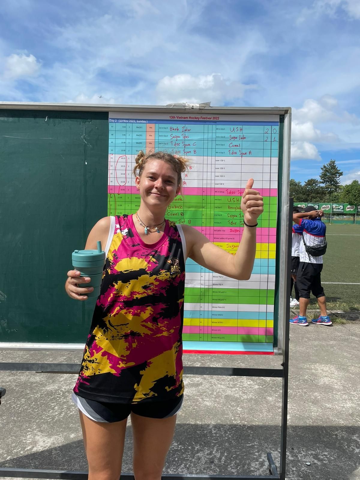 A woman is standing in front of a blackboard holding a cup and giving a thumbs up.