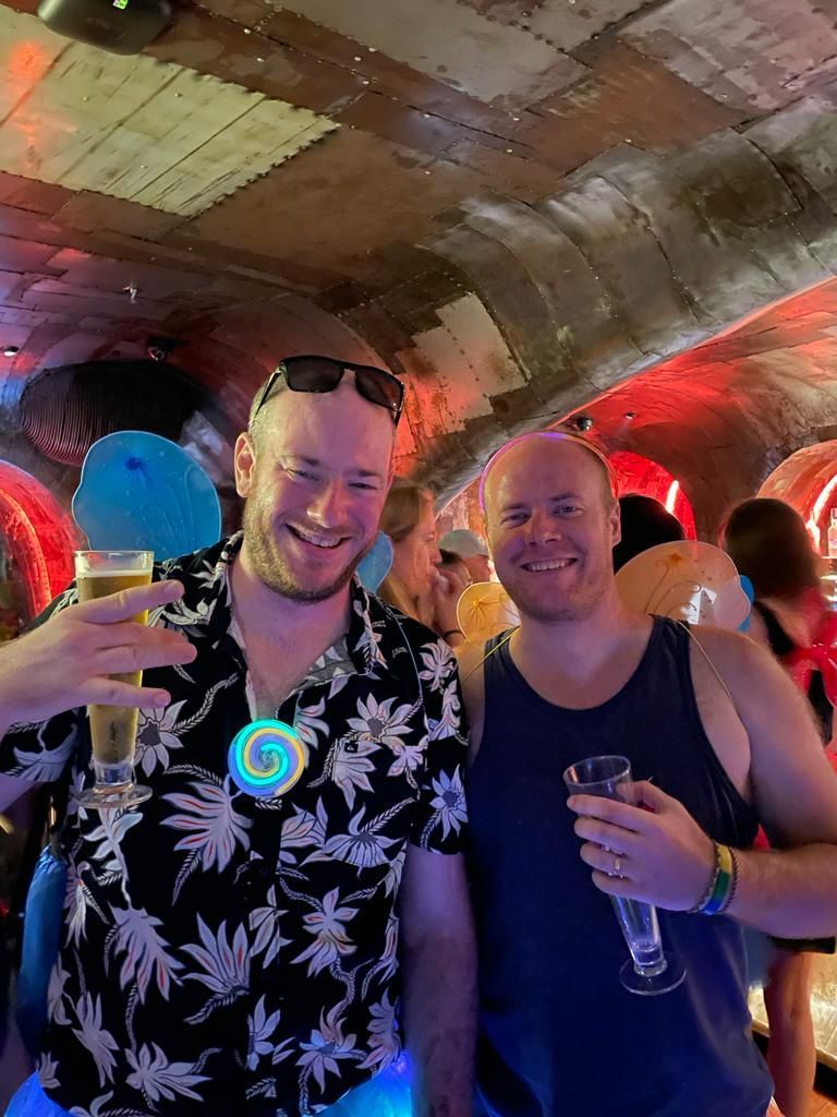 Two men are standing next to each other in a dark room holding drinks.