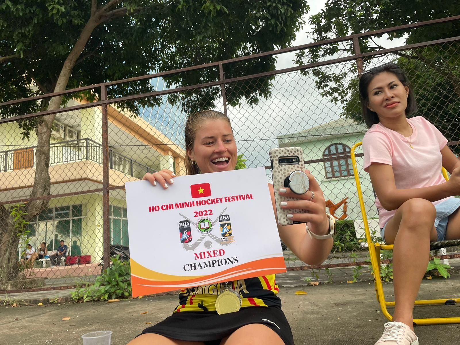 A woman is holding a sign and taking a selfie with her phone.