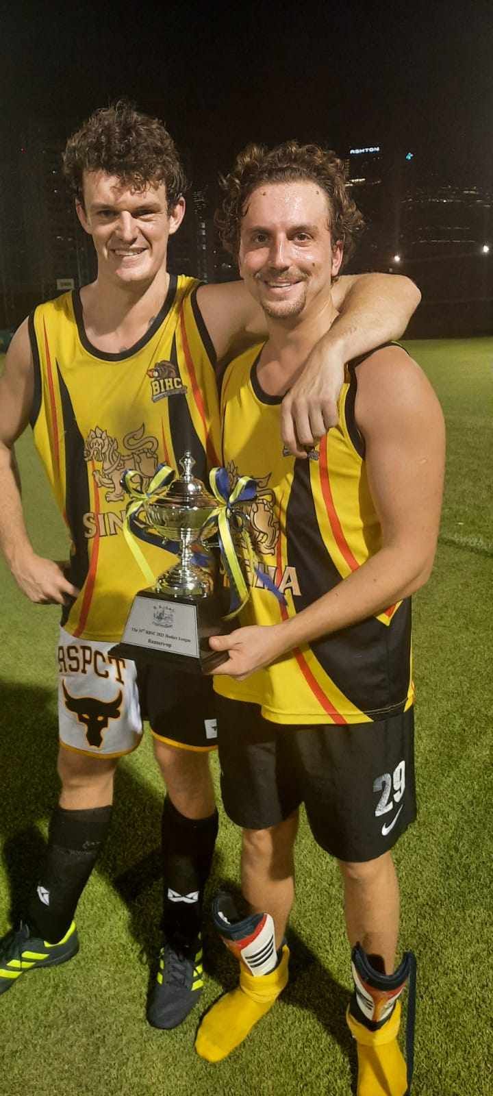 Two men are standing next to each other on a soccer field holding a trophy.
