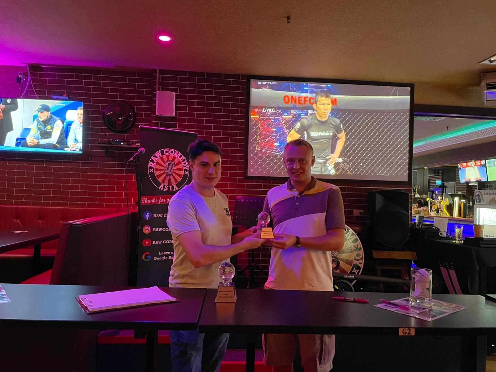 Two men are standing next to each other in a bar holding a trophy.