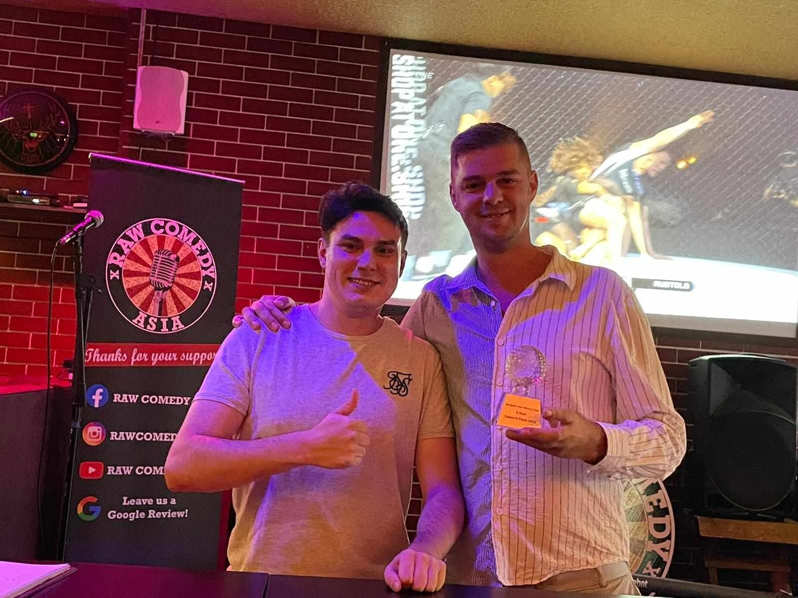 Two men are standing next to each other in a room holding a trophy and giving a thumbs up.